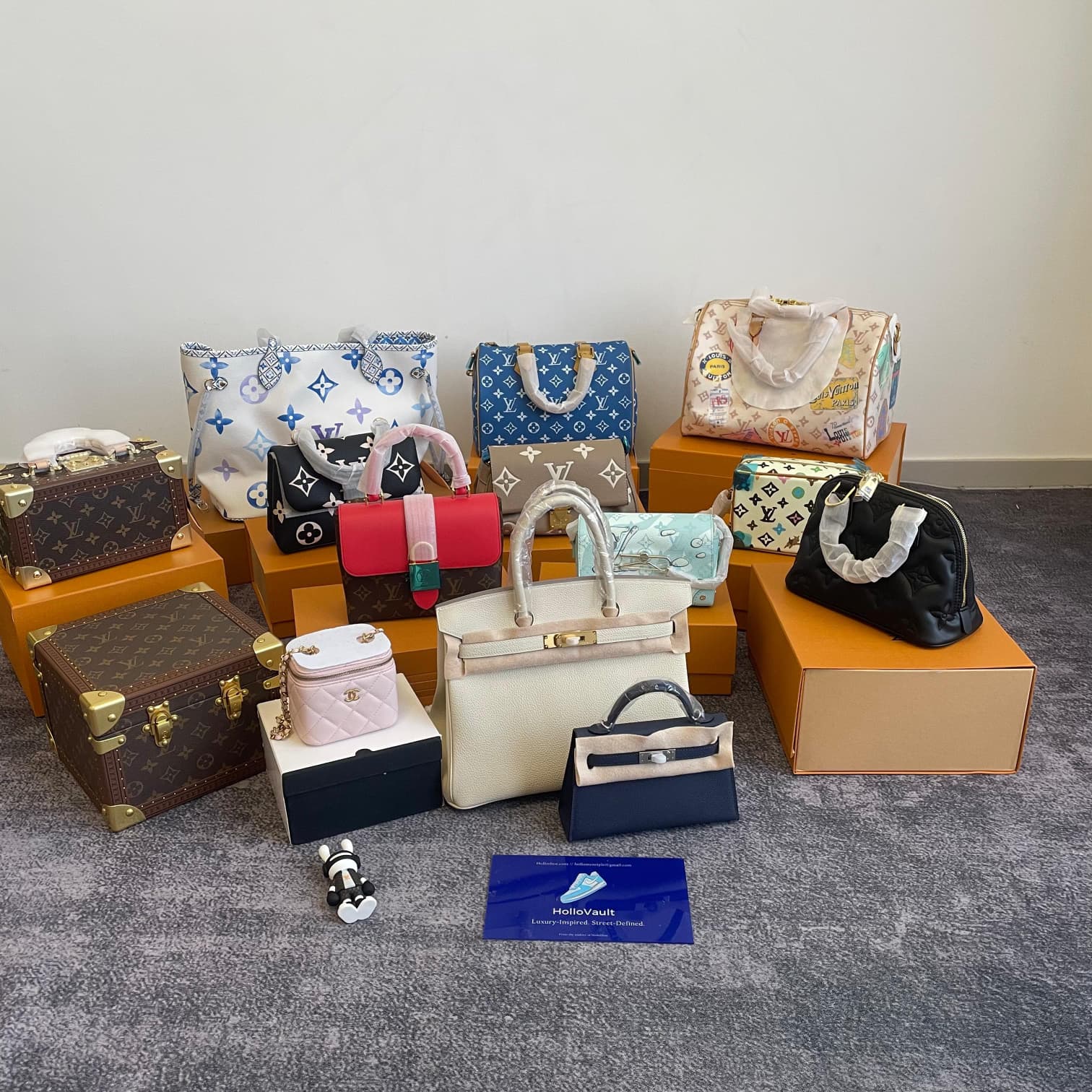 Various luxury designer bags from Louis Vuitton, displayed on orange boxes, including a mix of iconic styles such as mini trunks, handbags, and colorful bags with unique details. The bags are arranged neatly on a soft gray surface, showcasing their elegant design and craftsmanship.