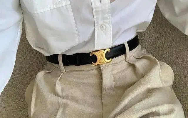 Close-up of a person wearing beige high-waisted trousers, a crisp white button-down shirt, and a black textured leather Celine Triomphe belt with a prominent gold-tone hardware buckle.