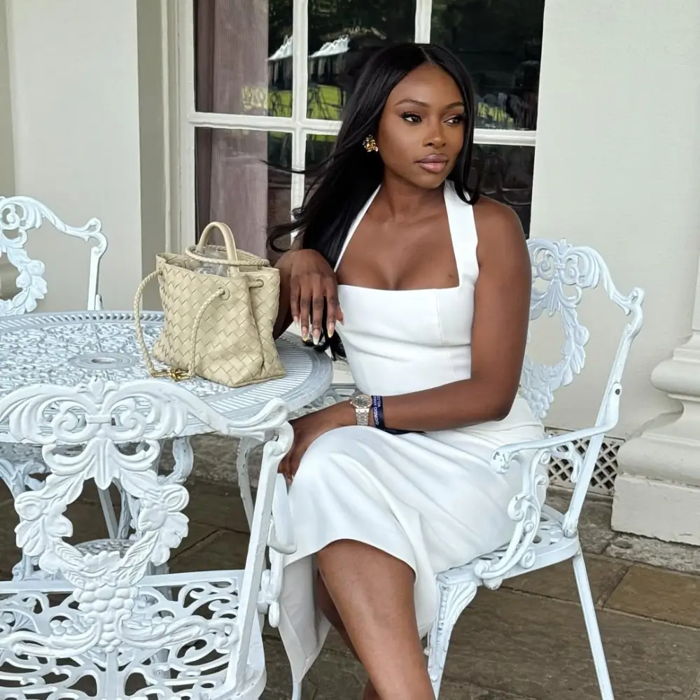 Elegant Black woman sitting outdoors in a white halter midi dress paired with a woven cream luxury bucket bag for a chic summer brunch outfit inspiration.