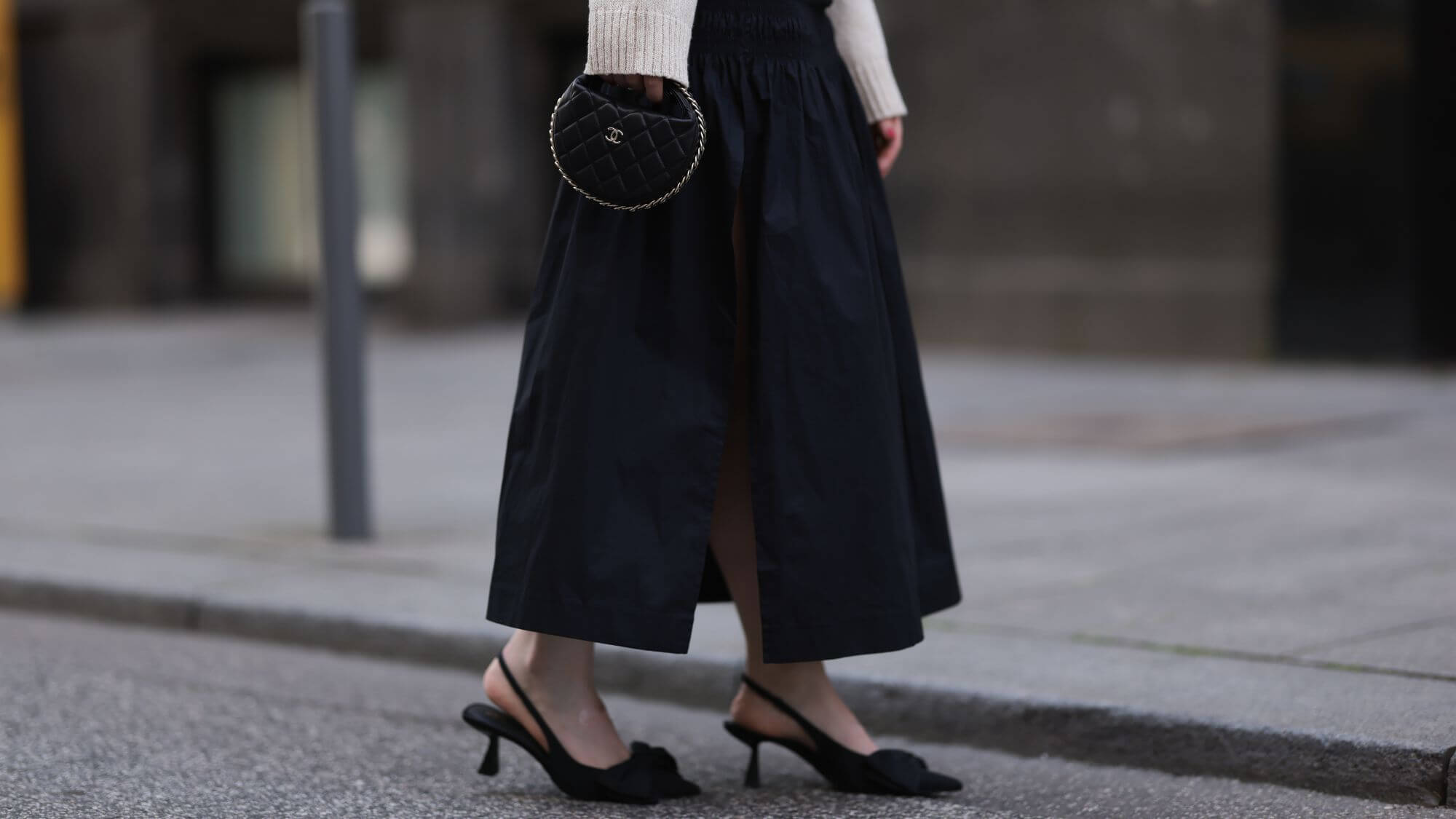 Close-up street style outfit featuring a woman in a black side-slit midi skirt, black pointed-toe slingback heels, and a quilted Chanel heart clutch bag.